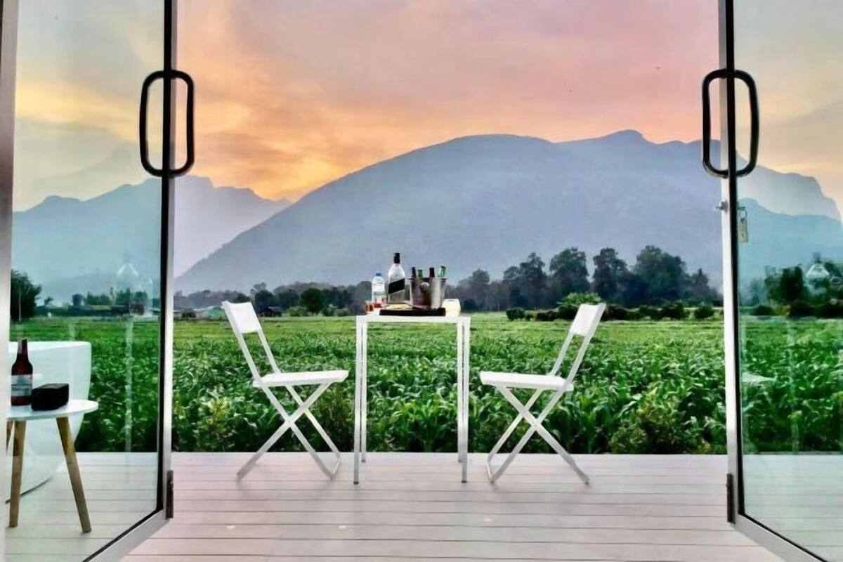 A room with a view of a green field and mountains. There is a table with two chairs and a bottle on it.