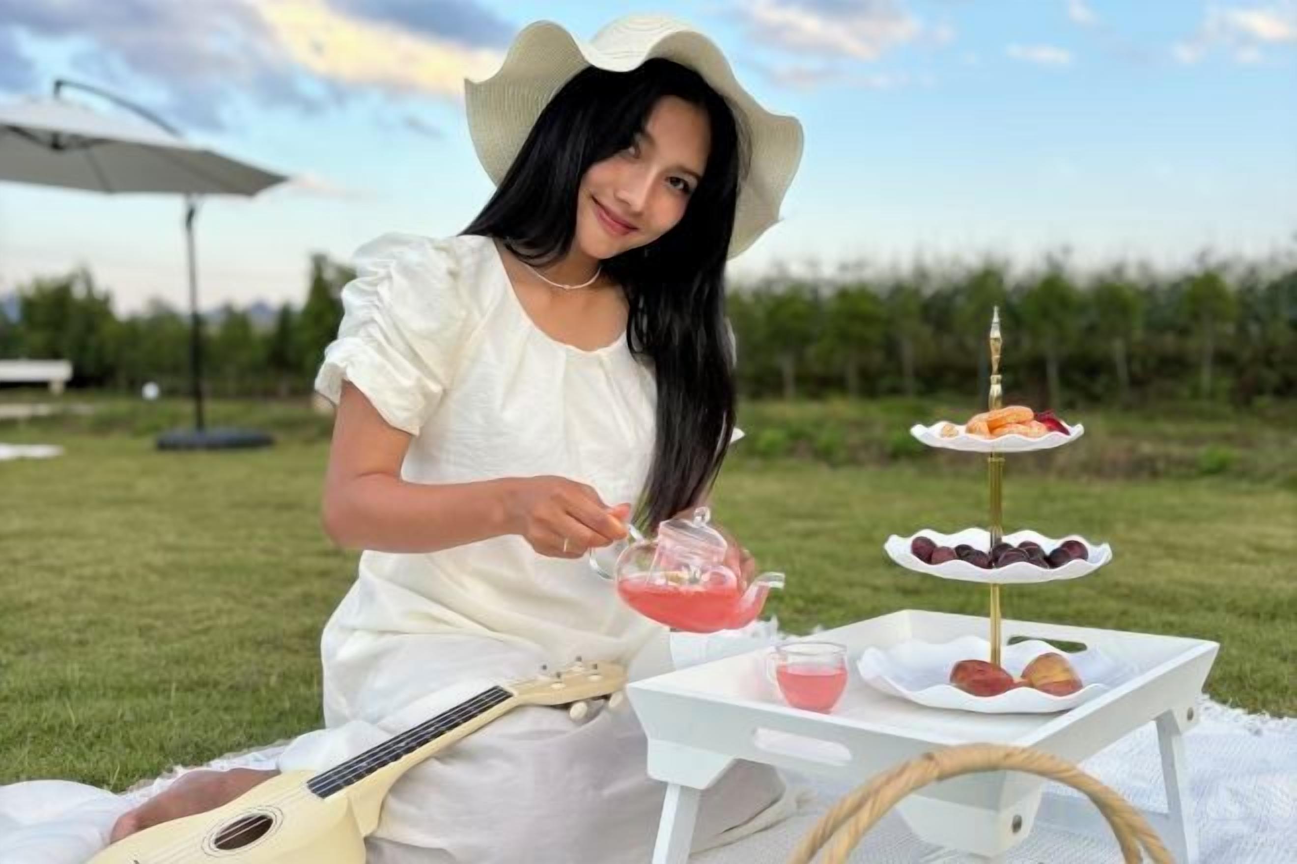 A person sitting outdoors with a guitar, a tray with desserts, and a drink.