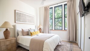 Ein Schlafzimmer mit einem Bett, zwei Nachttischen, einem großen Fenster mit Vorhängen, einem an der Wand befestigten Fernseher und einem gerahmten Kunstwerk.