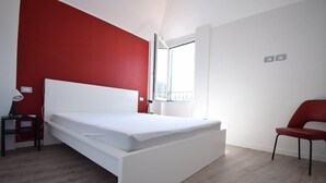 A modern bedroom with a white bed, red accent wall, and a window providing natural light. - Casa Luisa - Surrounded by greenery, just steps from the sea (Borganzo)