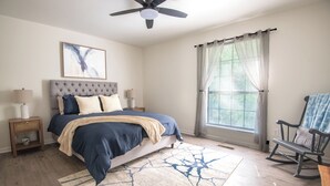 A bedroom with a bed, a ceiling fan, a window with curtains, a rocking chair, and a bedside table with a lamp. - Maple & Marigold – Modern Comfort + WiFi (Birmingham)