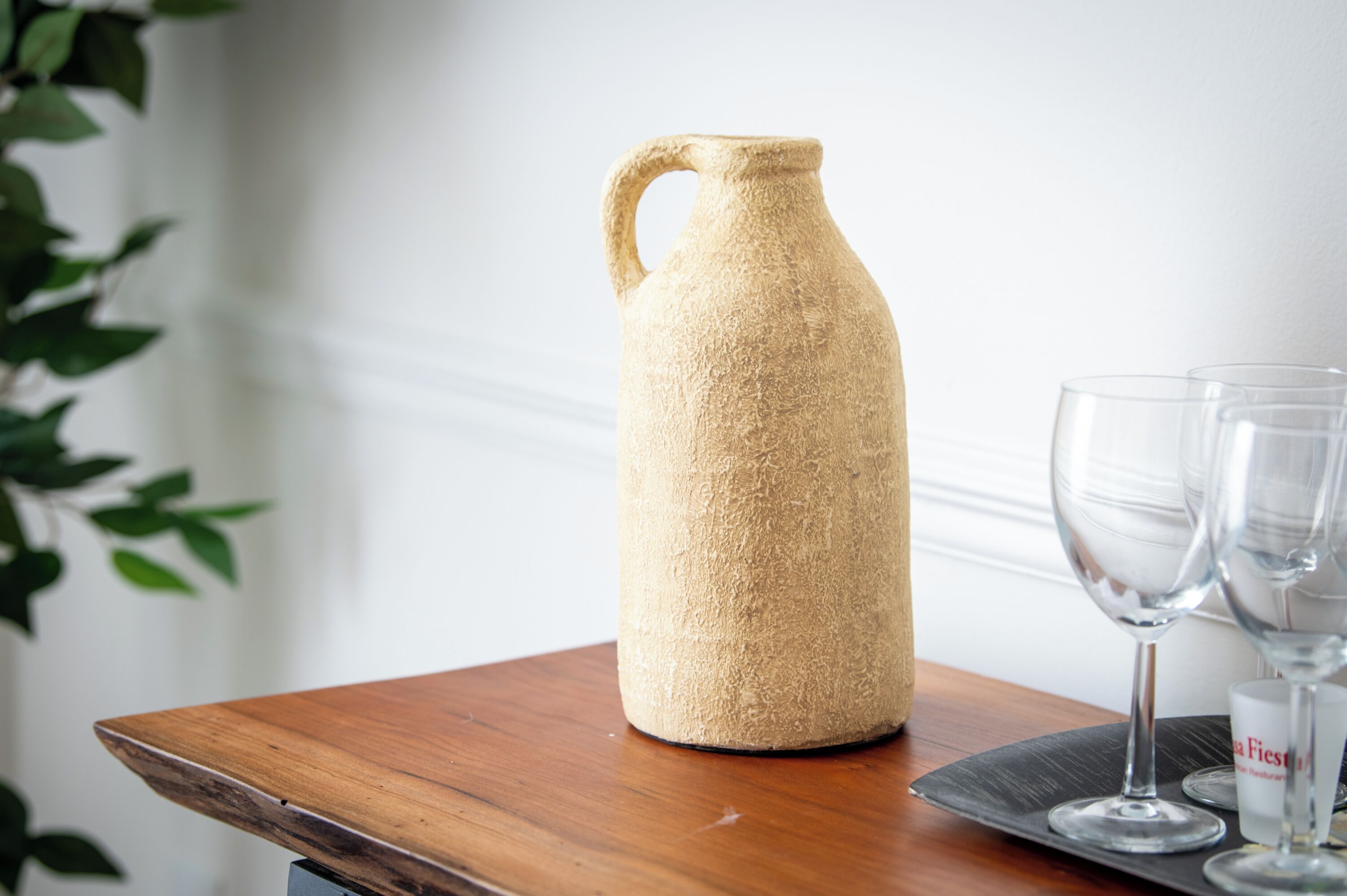 A textured, beige ceramic pitcher on a wooden table with two clear wine glasses beside it.