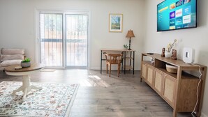 A living room with a TV displaying streaming services, a wooden cabinet, a table with a chair, and a large window with blinds. - Maple & Marigold – Modern Comfort + WiFi (Birmingham)