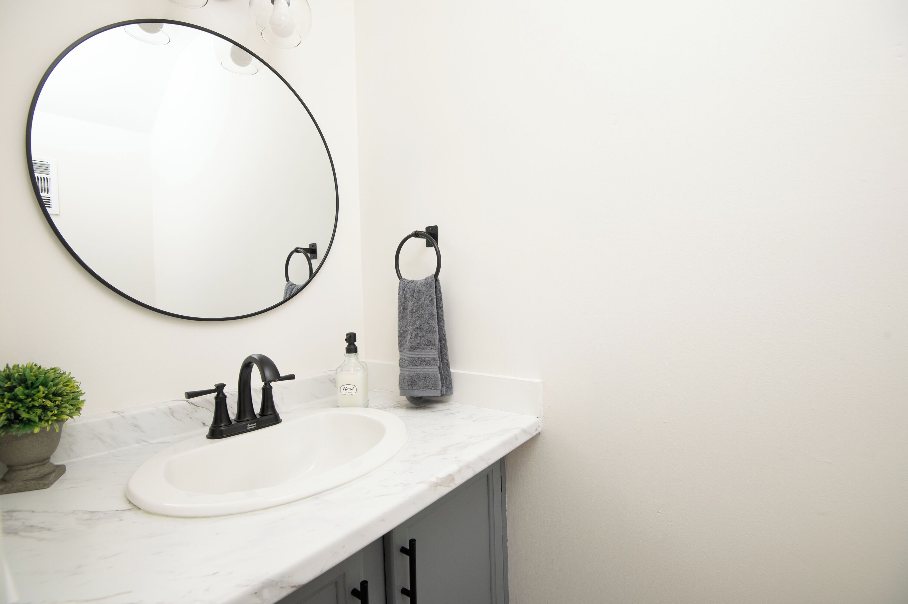 A bathroom with a round mirror, a white sink, a black faucet, a towel, a soap dispenser, and a small potted plant.