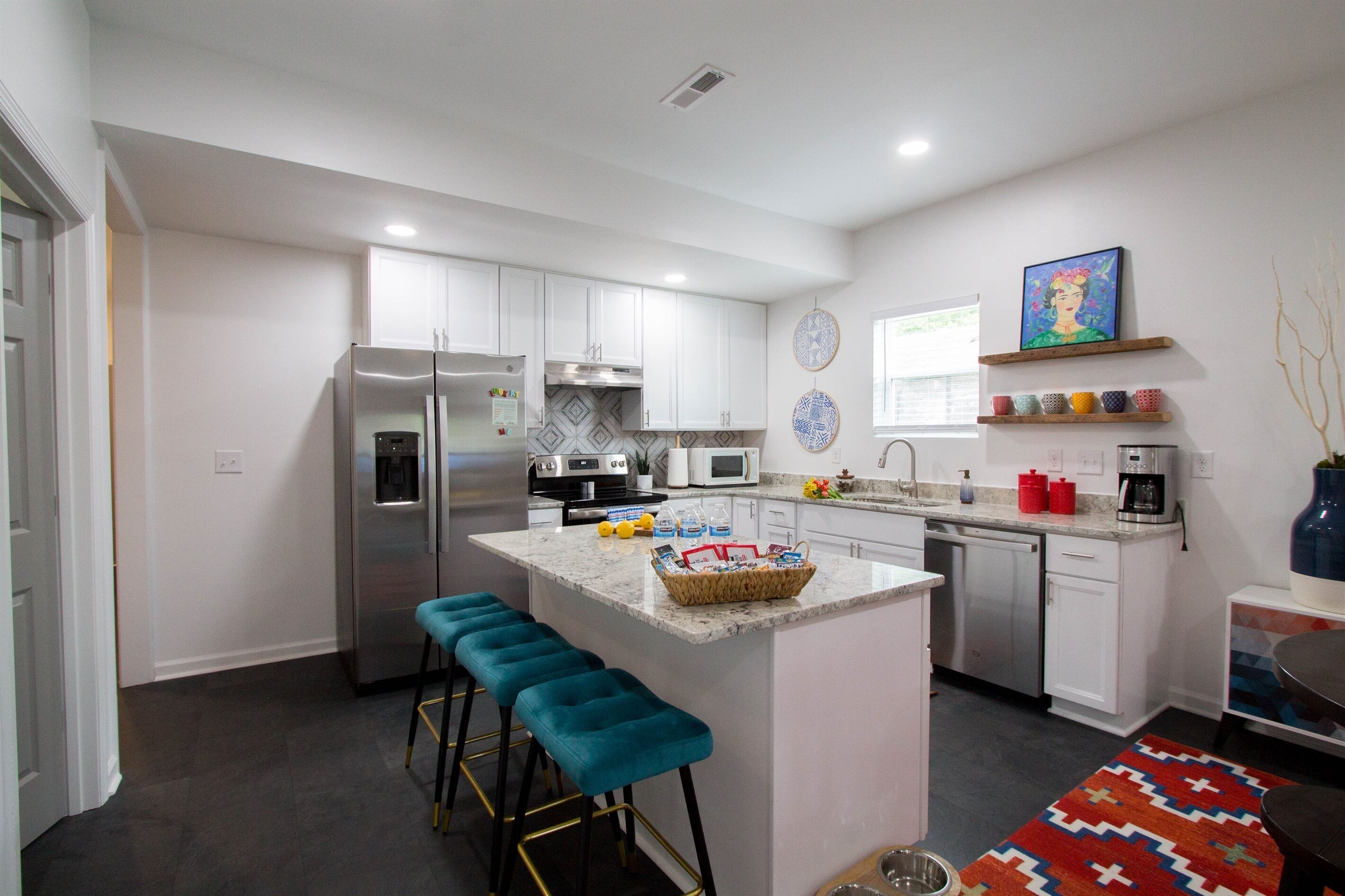 A modern kitchen with white cabinets, a stainless steel refrigerator, a microwave, and a coffee maker. There are blue bar stools and a colorful rug.