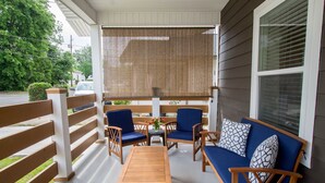 Uma área de estar coberta ao ar livre, com uma mesa de madeira, cadeiras azuis com almofadas e um sofá com coussins estampados.