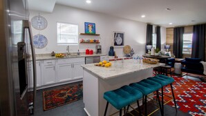 Uma cozinha moderna com armários brancos, uma ilha central com banquetas e uma área de jantar com sofá azul.