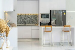 A modern kitchen with white cabinetry, a tiled backsplash, and a stainless steel refrigerator. There are two bar stools and a small island with a sink. - Residence Villa｜Modern 3 Bedroom Pool Villa｜Warm Plus (Phuket)