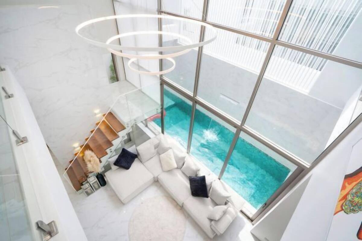 Un intérieur moderne avec une grande baie vitrée donnant sur une piscine, un canapé blanc agrémenté de coussins et un luminaire circulaire.