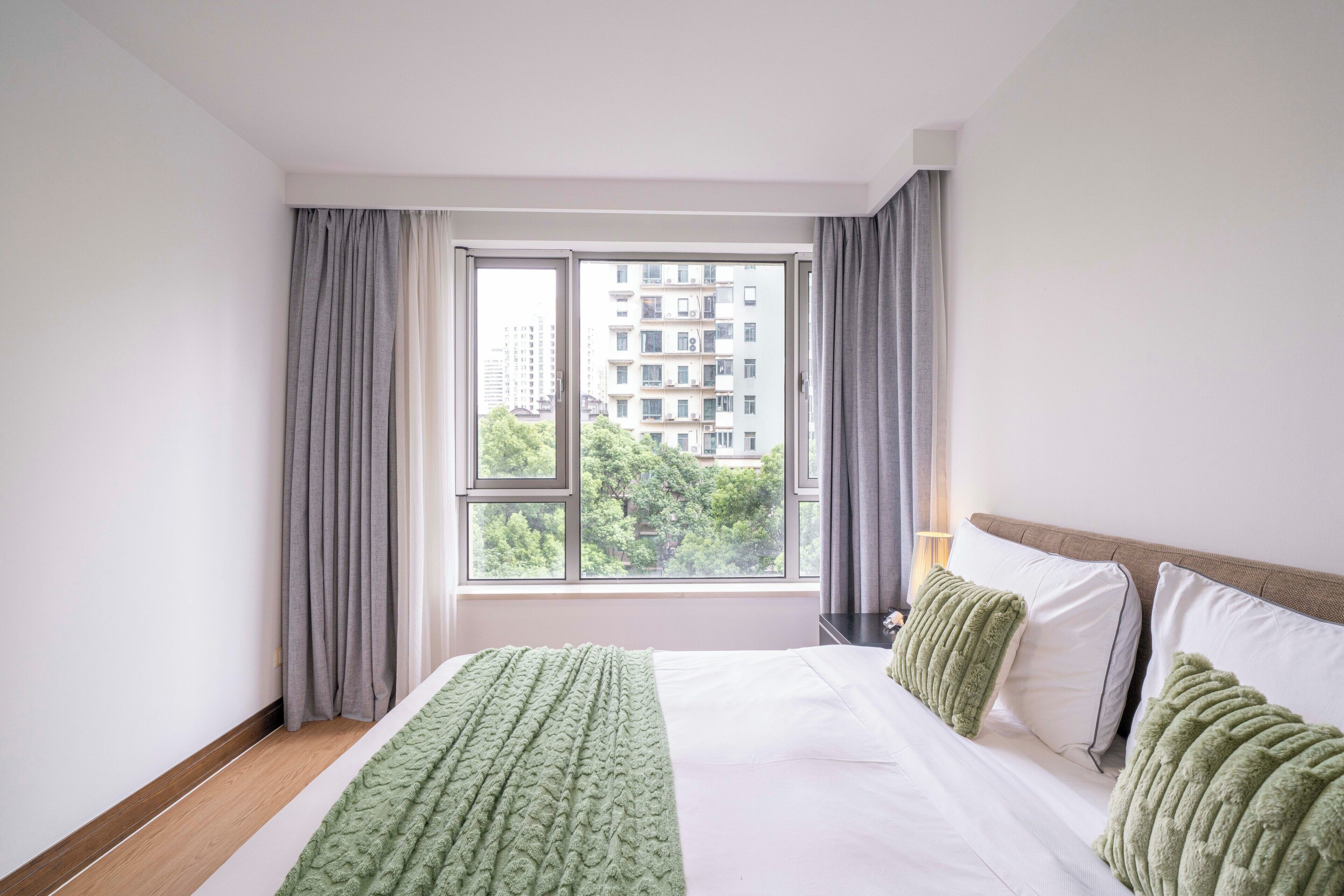 Una habitación de hotel con un ventanal grande, una cama con sábanas blancas y una manta verde.