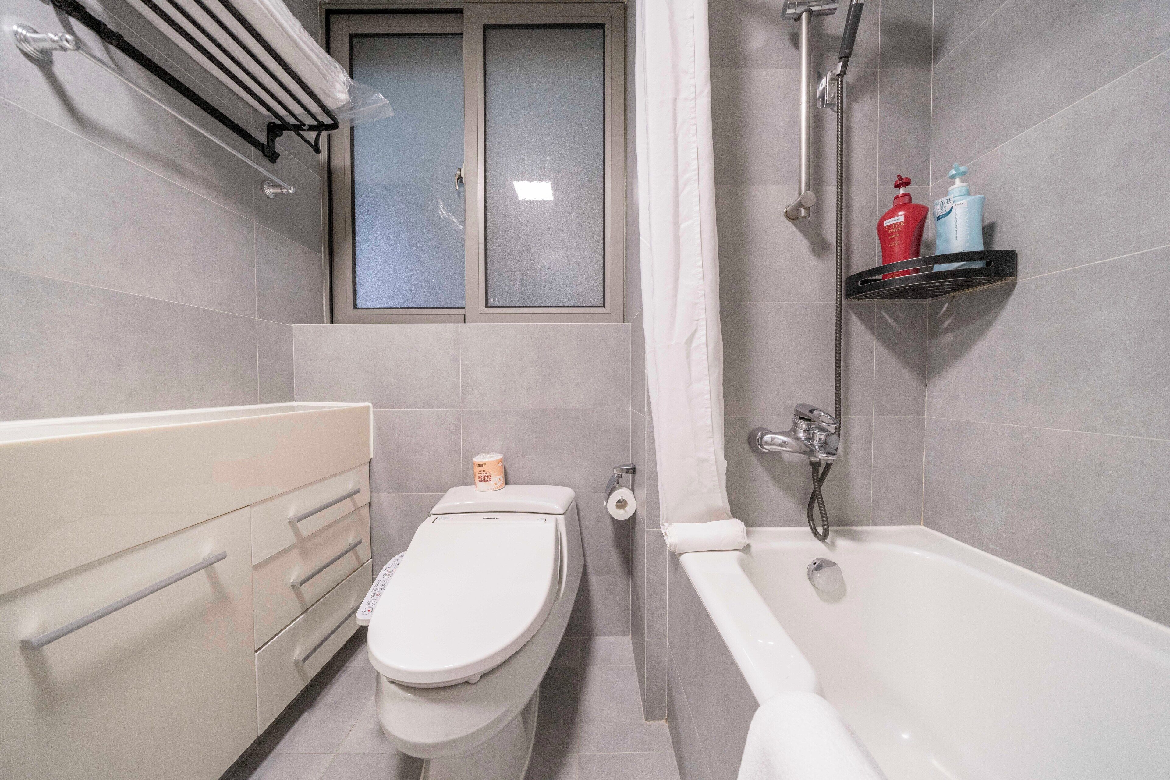 A bathroom with a toilet, a bathtub, a window, and a shelf with toiletries.