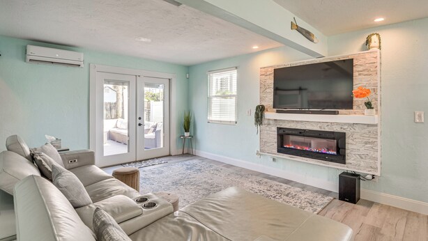 A living room with a beige sectional sofa, a flat-screen TV mounted above a fireplace, and a sliding glass door leading to an outdoor area. - Walk to Ocean! Home w/ Large Yard in Ormond Beach (Ormond Beach)