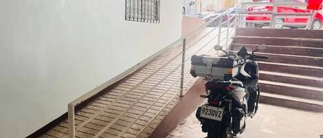 Un'area esterna coperta con una motocicletta parcheggiata a destra, una scala che conduce a un veicolo rosso e una ringhiera metallica lungo il lato sinistro.
