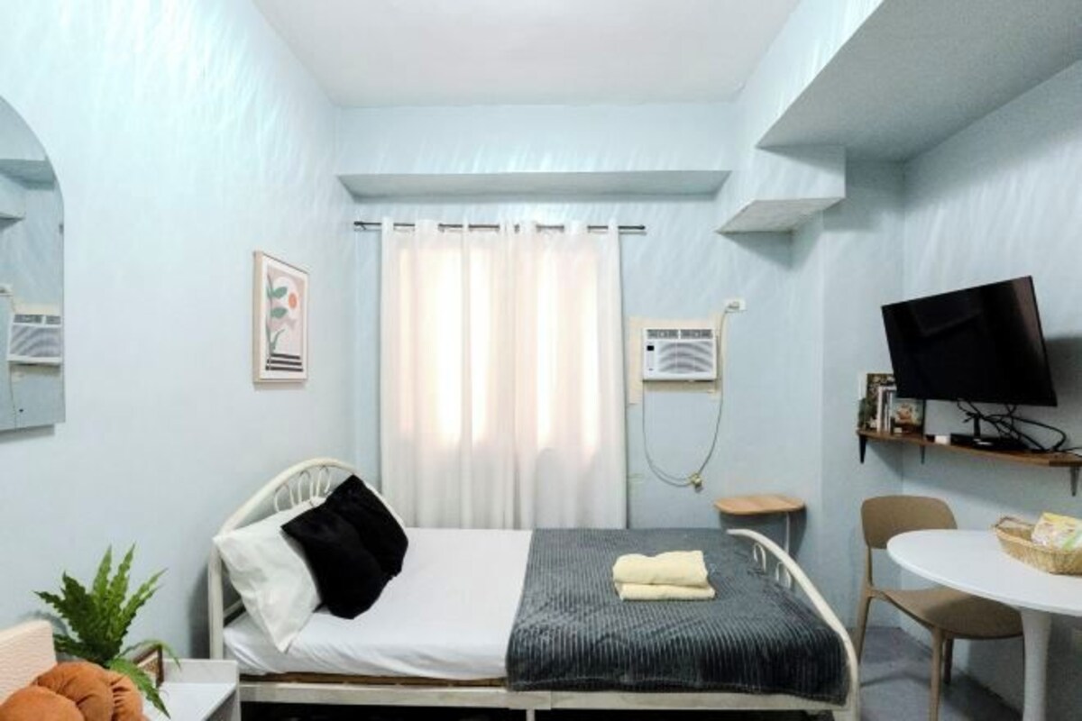 A bedroom with a bed, a desk, a chair, a television, and a window with curtains.
