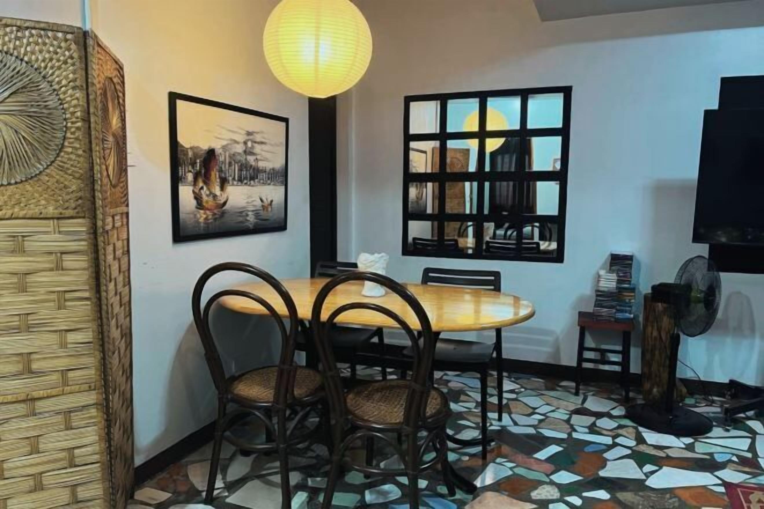 A dining area with a round table, chairs, a wall-mounted picture, a window with a view, and a ceiling fan.