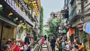 Un callejón estrecho con gente reunida alrededor de una vía de tren, rodeado de edificios con balcones y plantas colgantes.