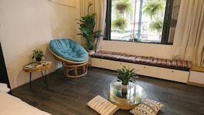 Una habitación acogedora con una cama, una silla azul, una mesita con una planta, un banco con cojines y un ventanal con plantas afuera.