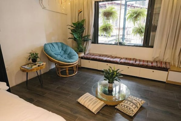A cozy room with a bed, a blue chair, a small table with a plant, a bench with cushions, and a large window with plants outside. - OH Trendy Homes Hoan Kiem LPH (Hanoi)