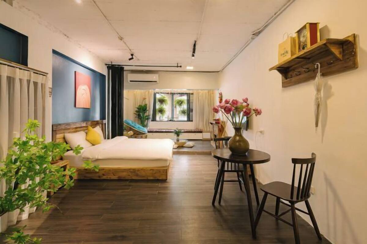 A room with a bed, a small table with chairs, a shelf with items, and a window with curtains.