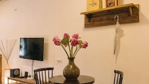 Un comedor con una mesa redonda, sillas, un florero con flores, un televisor y una repisa de madera con objetos.