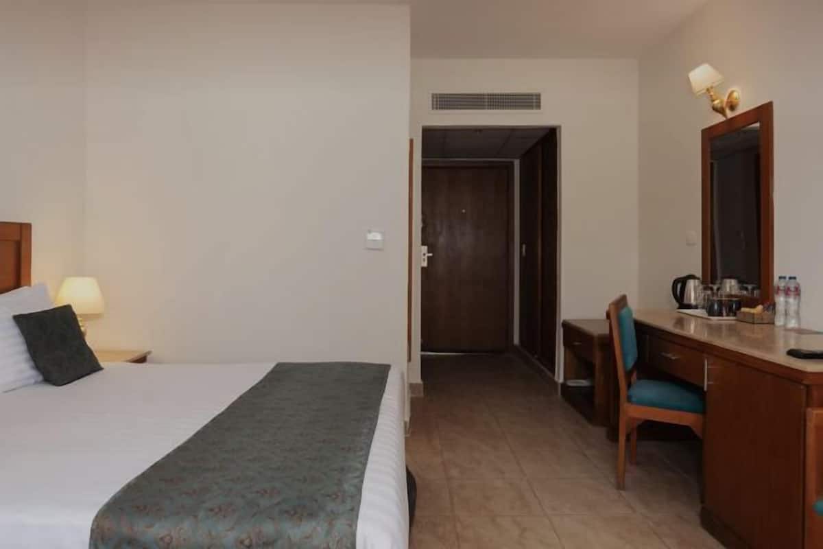 A hotel room with a bed, a desk, a chair, a mirror, and a kitchenette area.