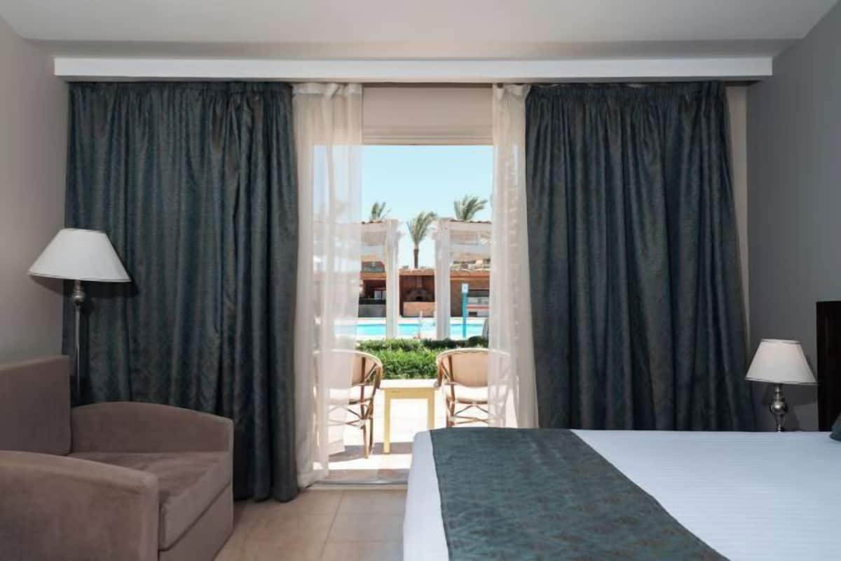A hotel room with a bed, two chairs, a lamp, and a view of a pool and palm trees.