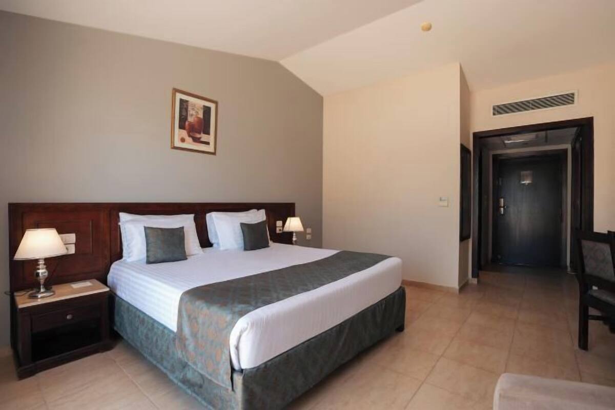 A hotel room with a large bed, two bedside tables with lamps, a framed picture on the wall, and a door leading to another room.