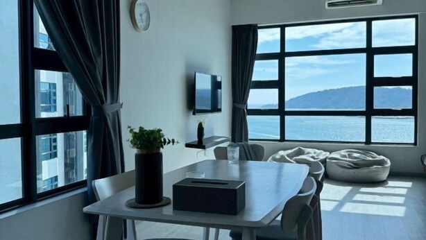 Une chambre moderne dotée d’une grande fenêtre donnant sur la mer. On y trouve une table avec une plante et une boîte, un téléviseur à écran plat fixé au mur, ainsi qu’un coin salon confortable avec un fauteuil poire.