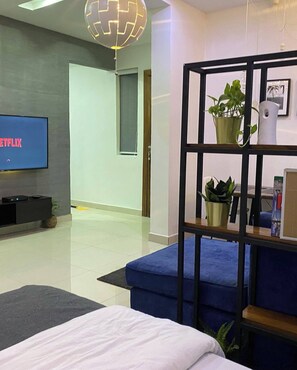 Een moderne hotelkamer met een grote flatscreen-tv waarop 'NETFLIX' staat, een zwarte metalen plank met planten, een blauwe bank en een bed met witte beddengoed.