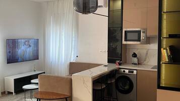 A modern living room with a flat-screen TV, a sofa, a dining table, and a kitchen area with a microwave and washing machine.
