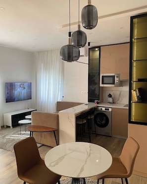 A modern living room with a sofa, a coffee table, and a TV. There is a kitchen area with a microwave and a washing machine.