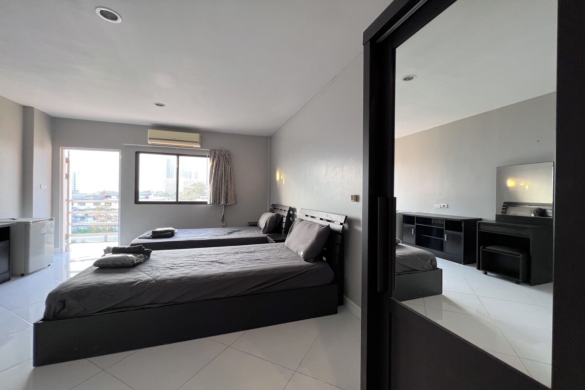 A modern hotel room with two beds, a kitchenette, and a large window providing a view of the city.