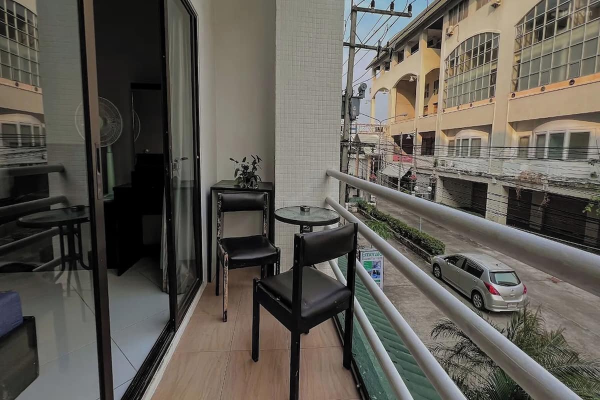 Un balcon avec une table et des chaises, une petite table avec une plante, et une vue sur une rue bordée d’immeubles et où l’on aperçoit une voiture.