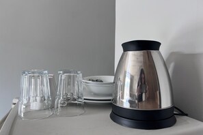 Une bouilloire en acier inoxydable posée sur un plan de travail blanc, à côté de deux verres transparents et d’un bol blanc.