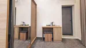 Habitación de hotel compacta con armario espejado, dos escritorios de madera con taburetes, un ventanal con persianas y aire acondicionado de pared.
