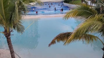 Zona de piscina con palmeras, una playa arenosa y gente nadando.