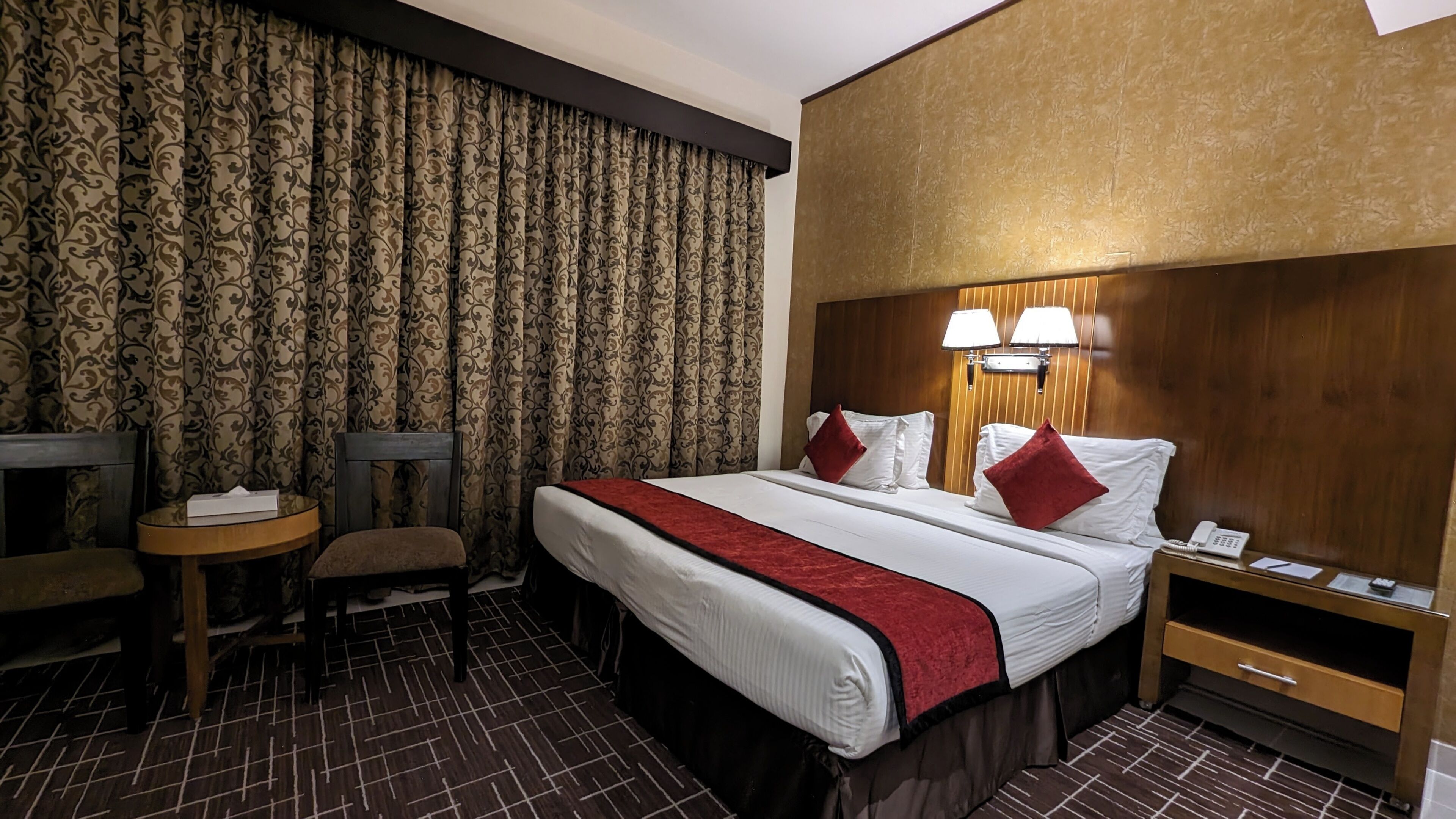 A hotel room with a large bed, two bedside lamps, a wooden headboard, a small table with a chair, and a telephone on the nightstand.