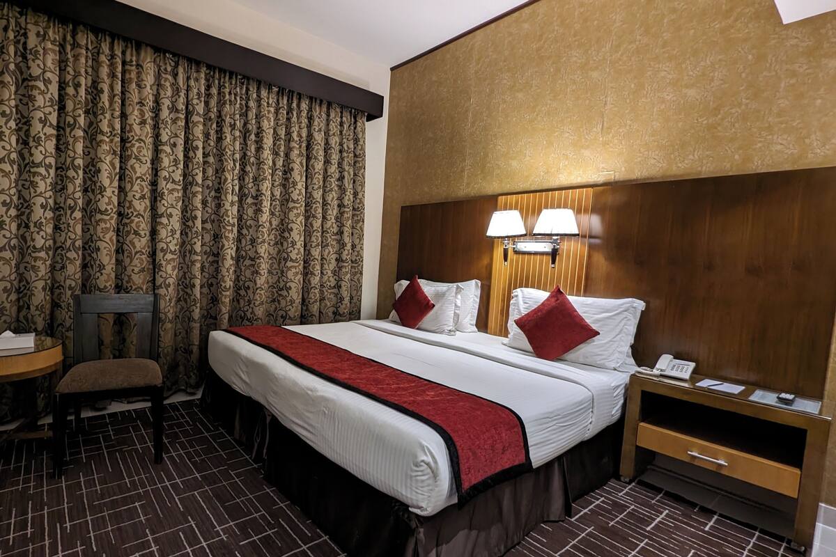 Habitación de hotel con una cama grande, dos lámparas de noche, un cabecero de madera, una mesita con una silla y un teléfono en la mesita de noche.