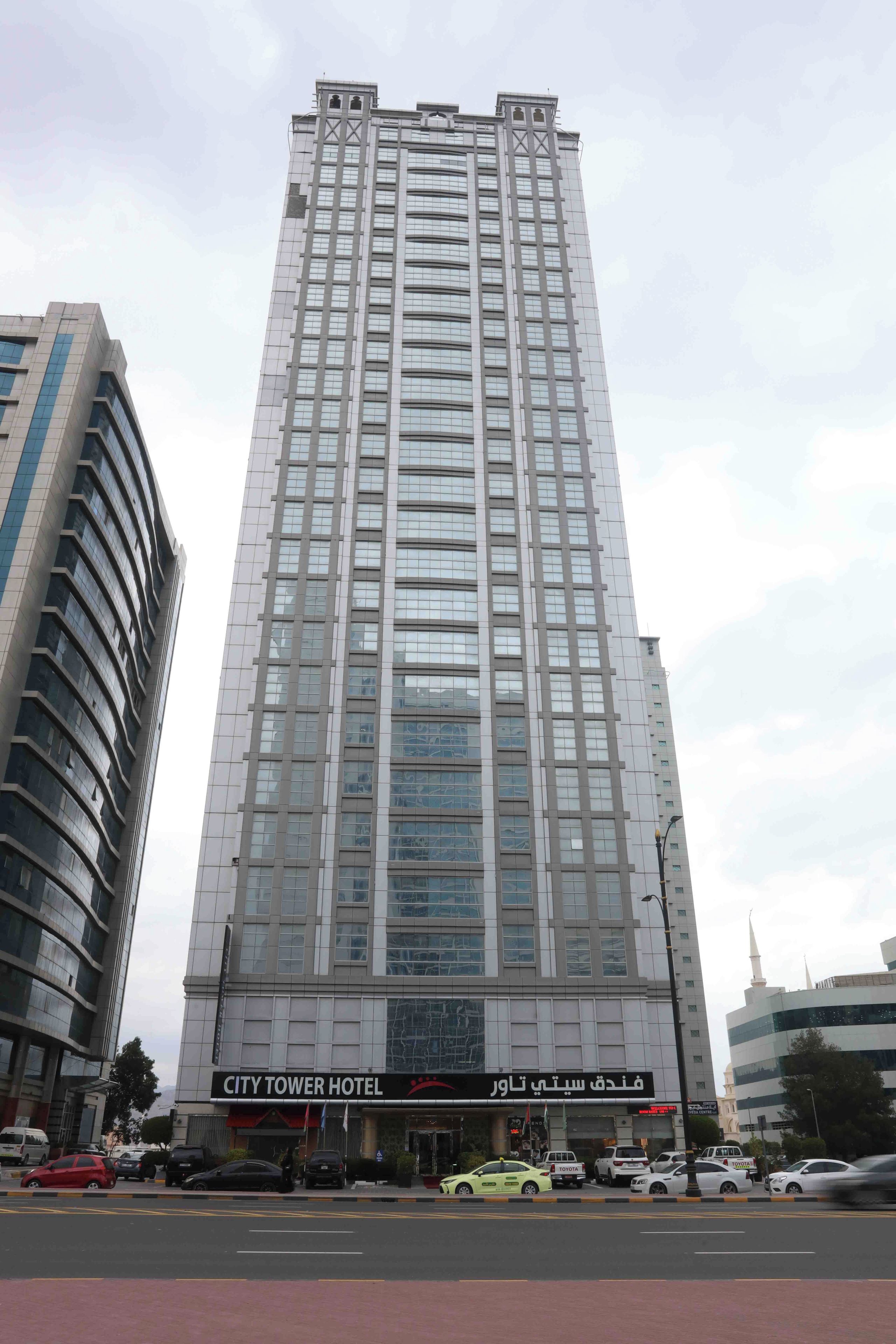 Een hoog hotelgebouw met een moderne gevel van glas, een opvallend bord met de tekst 'CITY TOWER HOTEL' en een drukke straat ervoor.