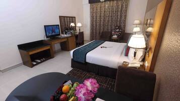 Une chambre d’hôtel comprenant un lit, un bureau avec une télévision, une table à manger avec des fruits et des fleurs, et un miroir.