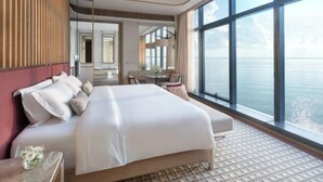 Une chambre d’hôtel avec un grand lit, un coin salon avec une table et des chaises, une grande fenêtre donnant sur la mer et un miroir au mur.