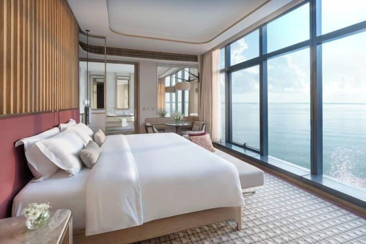 Habitación de hotel con una cama grande, una zona de estar con una mesa y sillas, un ventanal con vistas al mar y un espejo en la pared.