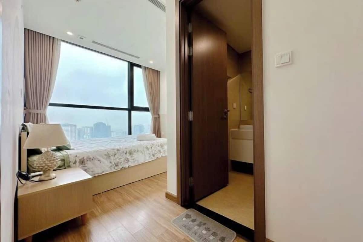 Quarto com cama, mesa de cabeceira, luminária e vista da cidade através de grandes janelas.