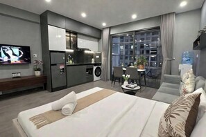 A modern hotel room with a bed, a small kitchenette, a dining area, and a large window.