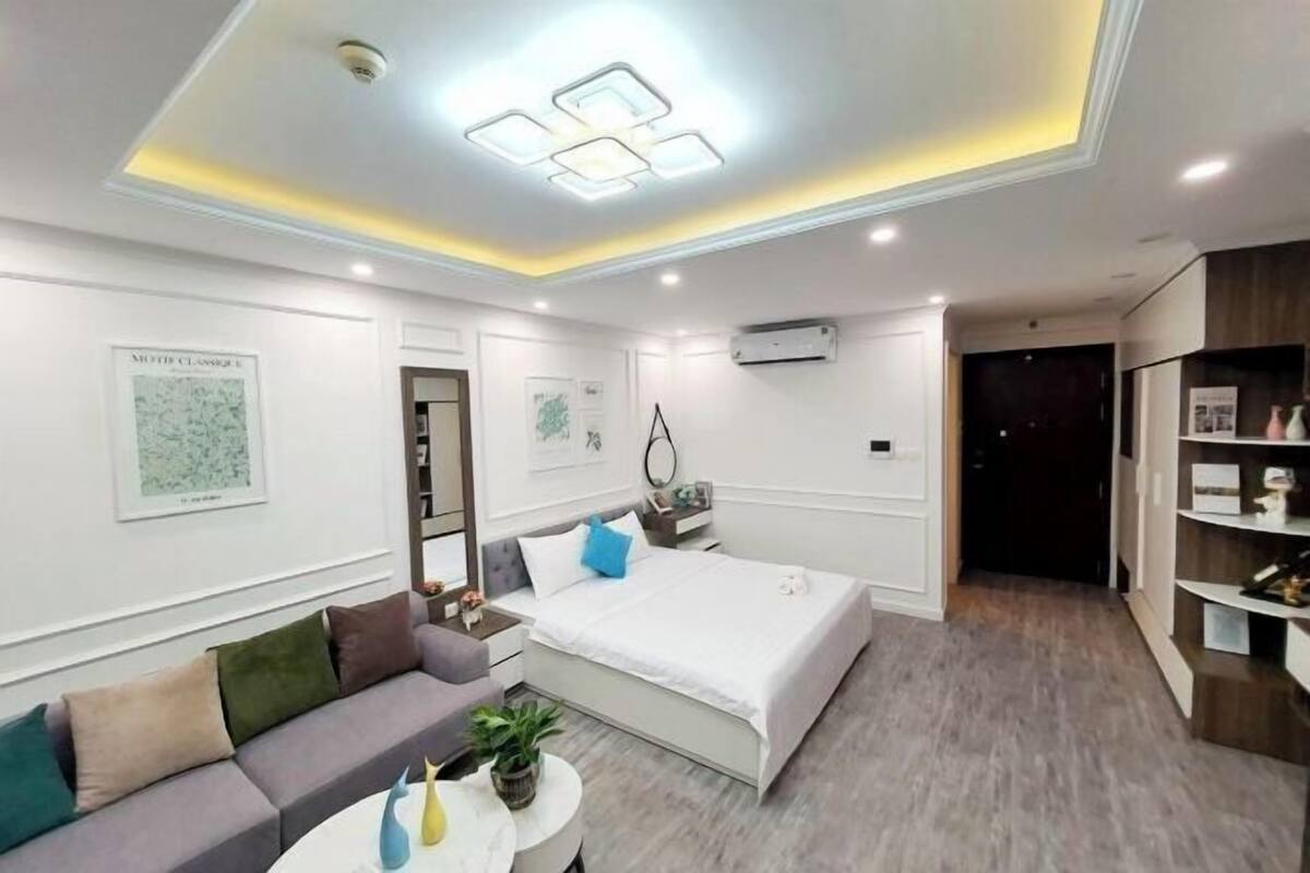 Quarto de hotel moderno com cama, sofá, uma mesinha com planta, luminária no teto e ar-condicionado fixado na parede.