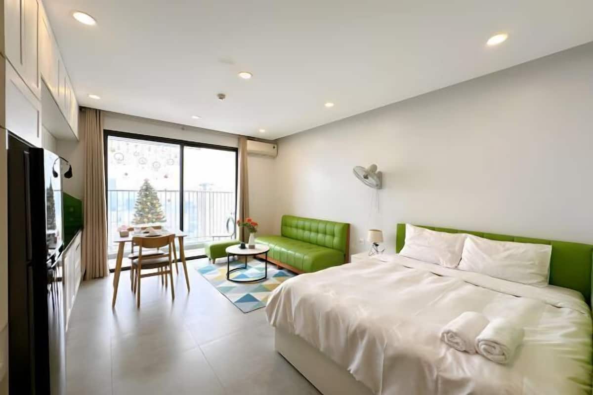 Quarto de hotel com cama, sofá verde, mesa de jantar com cadeiras, mesa redonda pequena com um vaso e uma árvore de Natal.