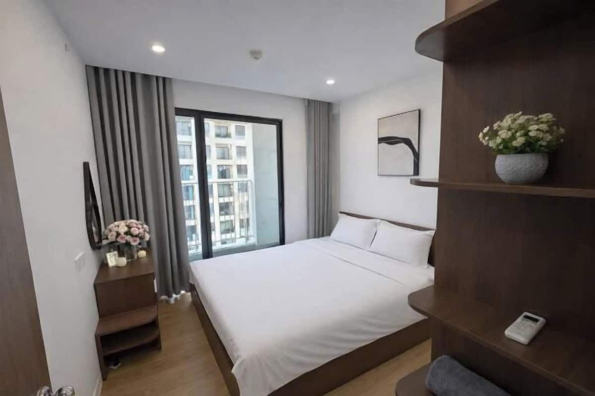 Quarto de hotel com cama, mesa de cabeceira, espelho, janela com cortinas e vista para edifícios.
