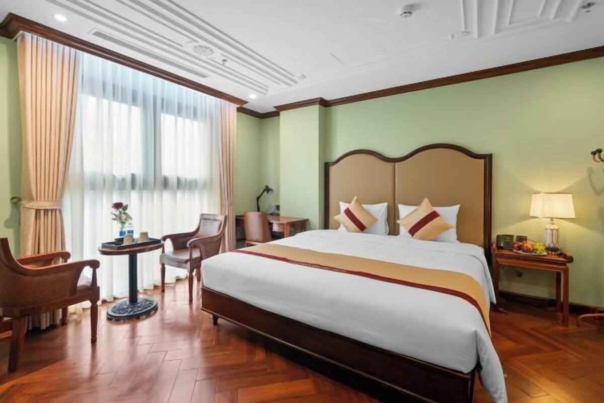 A hotel room with a large bed, a desk, a chair, a small table, and a lamp.