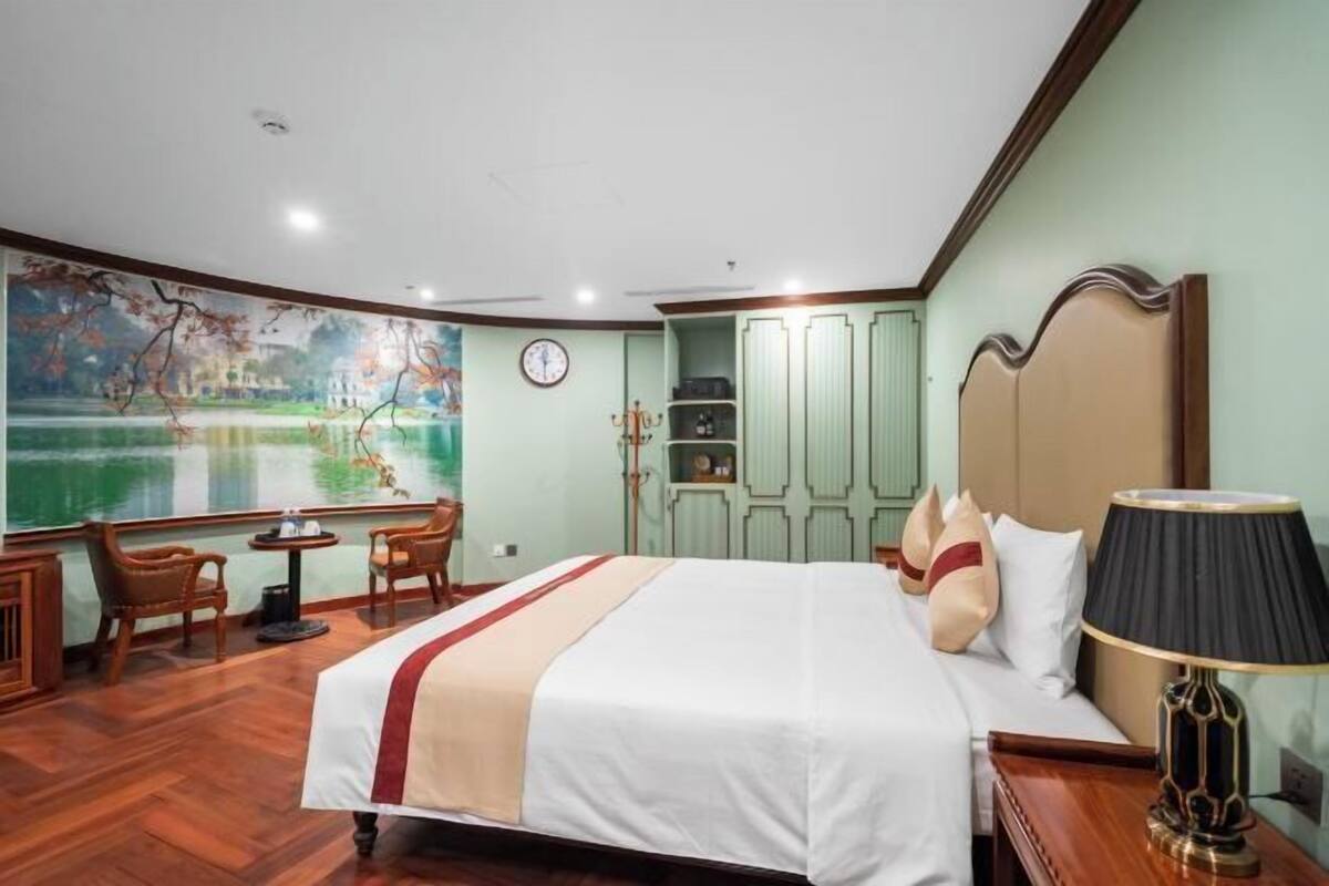 A hotel room with a large bed, a desk with a chair, a painting on the wall, and a view of a body of water.