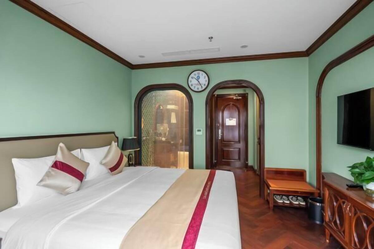 A hotel room with a bed, a clock, a television, and a wooden bench.